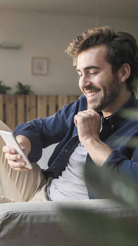 Mann mit Smartphone und lächelndem Gesichtsausdruck auf einem Sofa in einem Wohnzimmer.