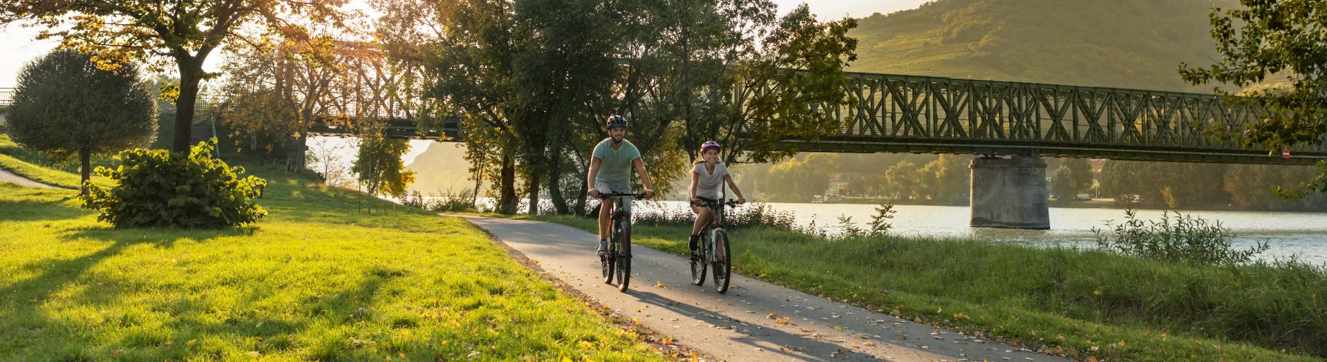 Zwei Personen radfahrend am Donauradweg bei Eisenbahnbrücke im Hintergrund