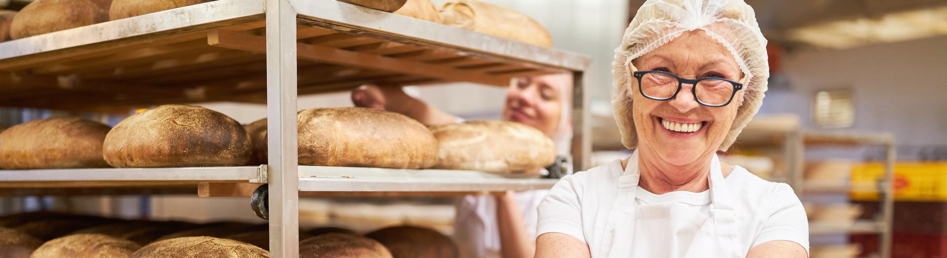Bäckerin steht in einer Backstube mit einem Laib Brot in der Hand und Backwaren im Hintergrund