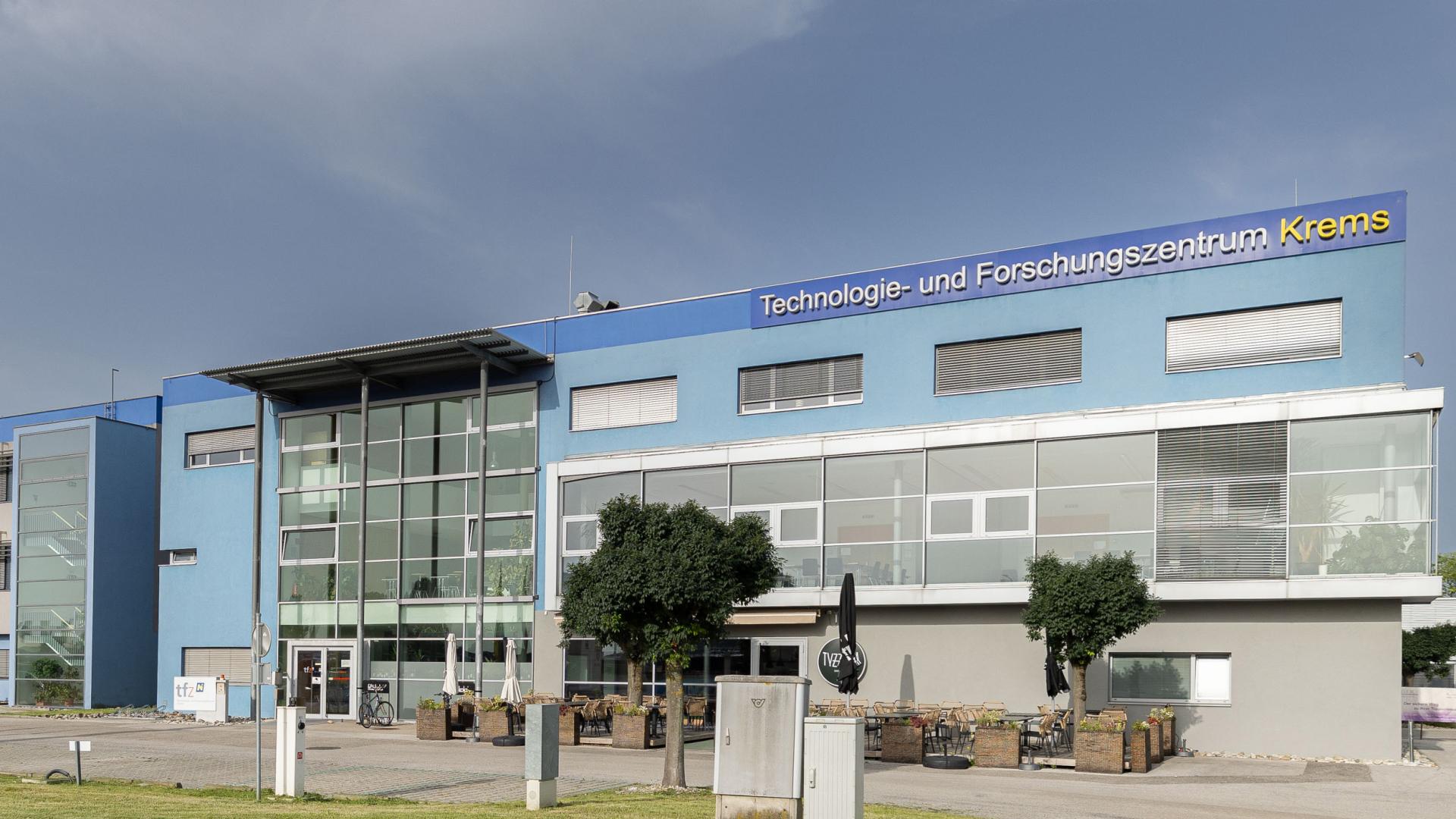 Teschnologie-und Fortschungszentrrum mit blauer Fassade, Parkplätzen, Gastrobereich und Pylone vor Haupteingang