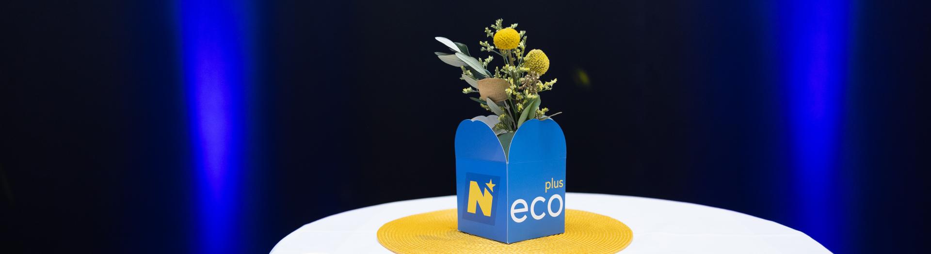 blauer ecoplus Kartonwürfel mit Blumenschmuck auf Stehtisch und blau beleuchteter Rückwand