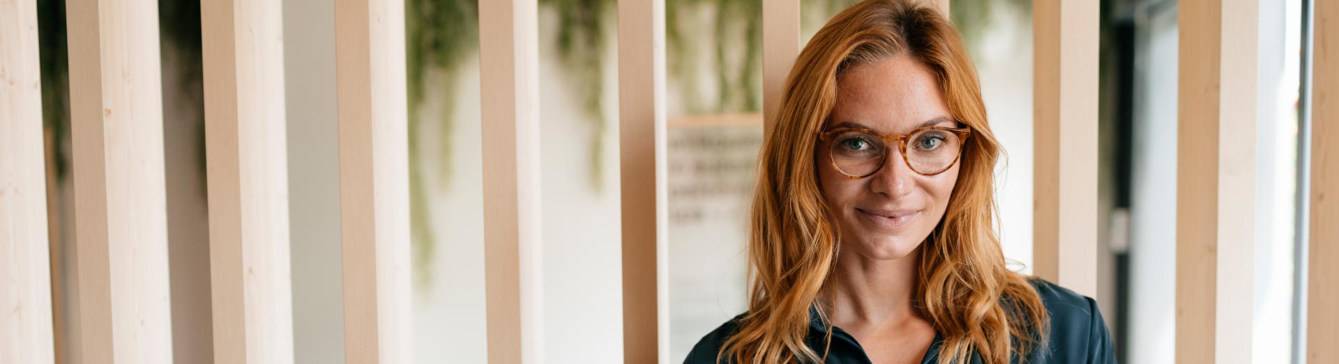 Frau mit Brille und verschränkten Armen vor einer Holztrennwand