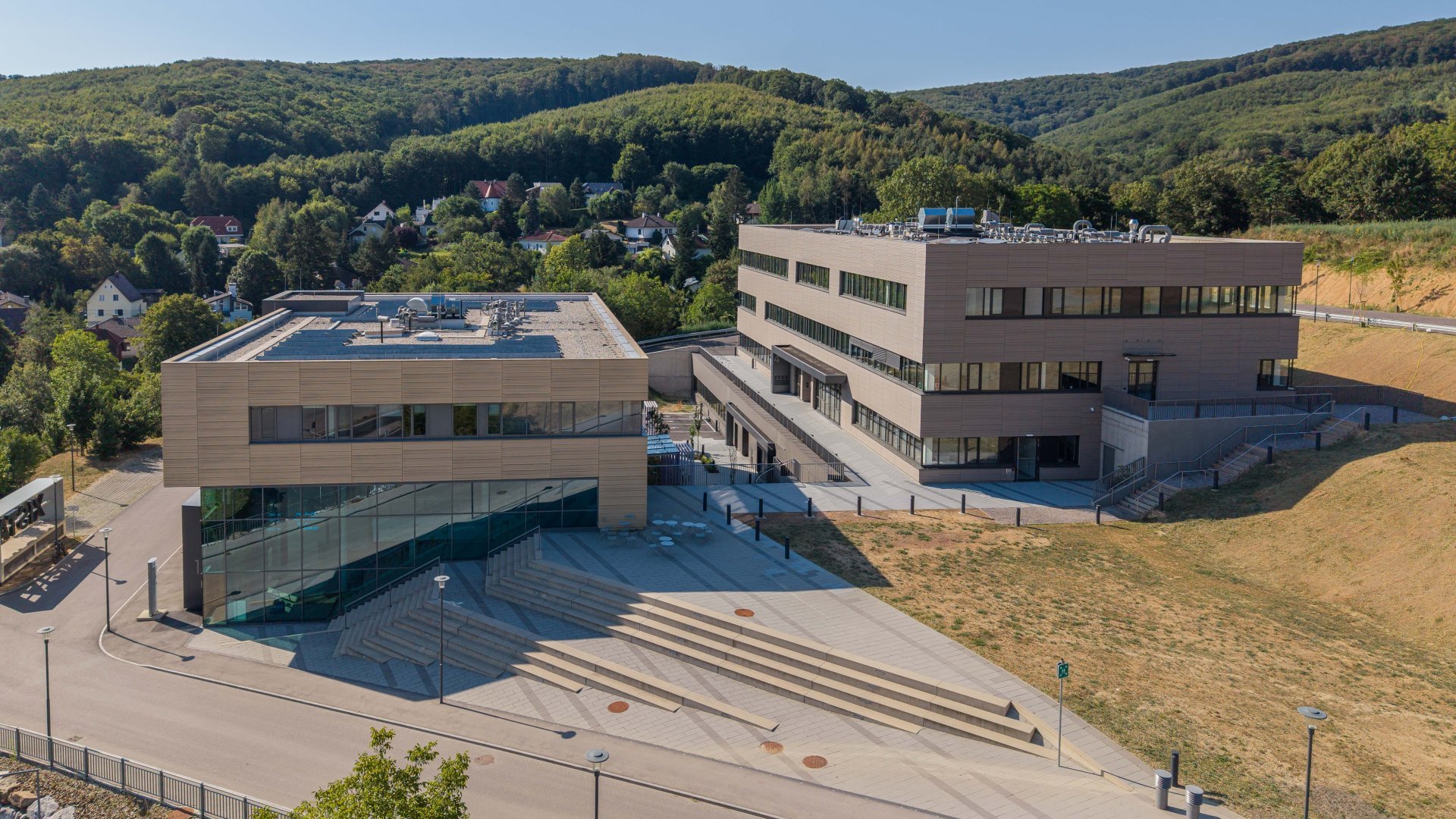 Drohnenansicht auf zwei Gebäude und großzügigen Außenstiegen vor einem großen Waldrücken