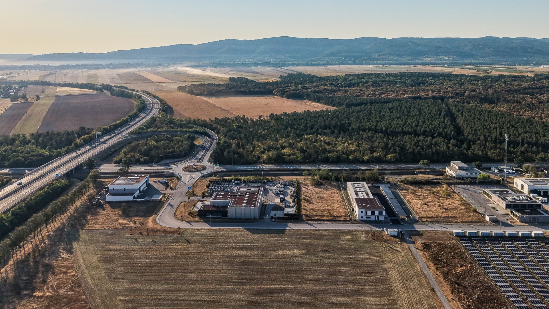 Drohnensicht über ein Betriebsgelände neben einer Schnellstraße, einem Kreisverkehr, Feldern und einem Föhrenwald
