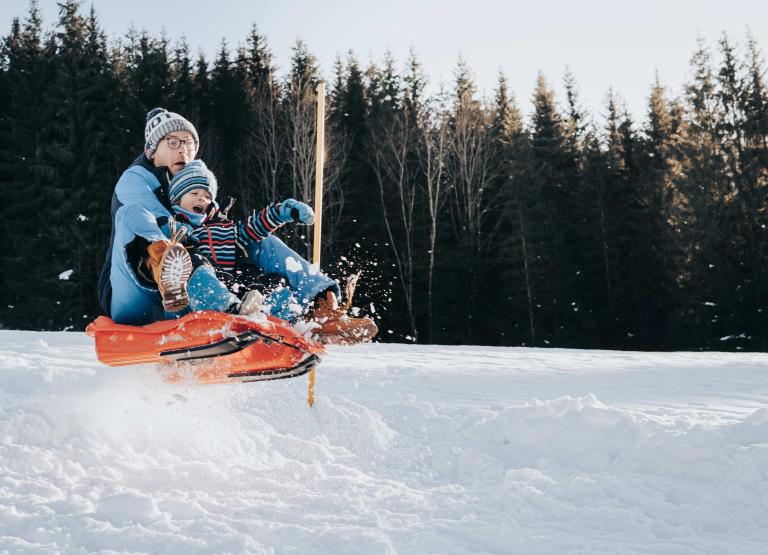 Mann mit kleinem Buben springen im kleinem Bob über Sprungschanze im Schnee