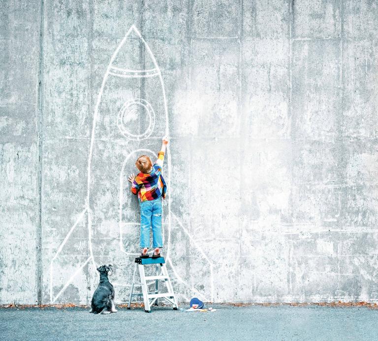 Kleiner Bub auf dreistufiger Leiter zeichnet mit Kreide Rakete an eine Wand