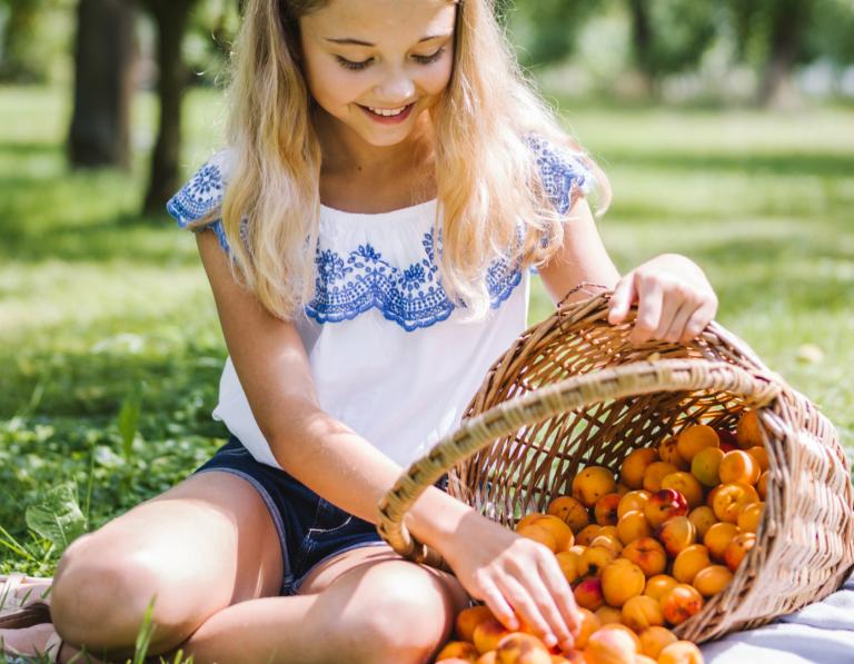 Mädchen sitzt auf Picknickdecke mit großen Korb voller Marillen