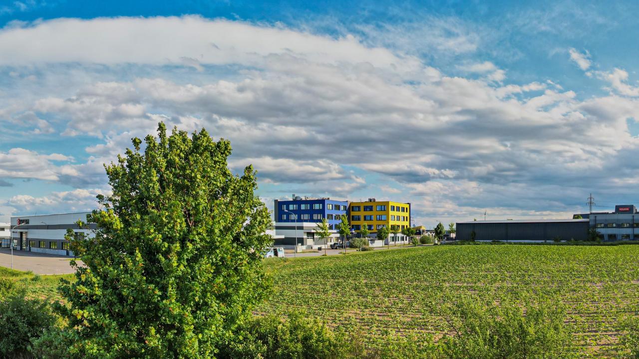 blau-gelber Gebäudekomplex mit Hallen hinter einem Windschutzgürtel mit Feld