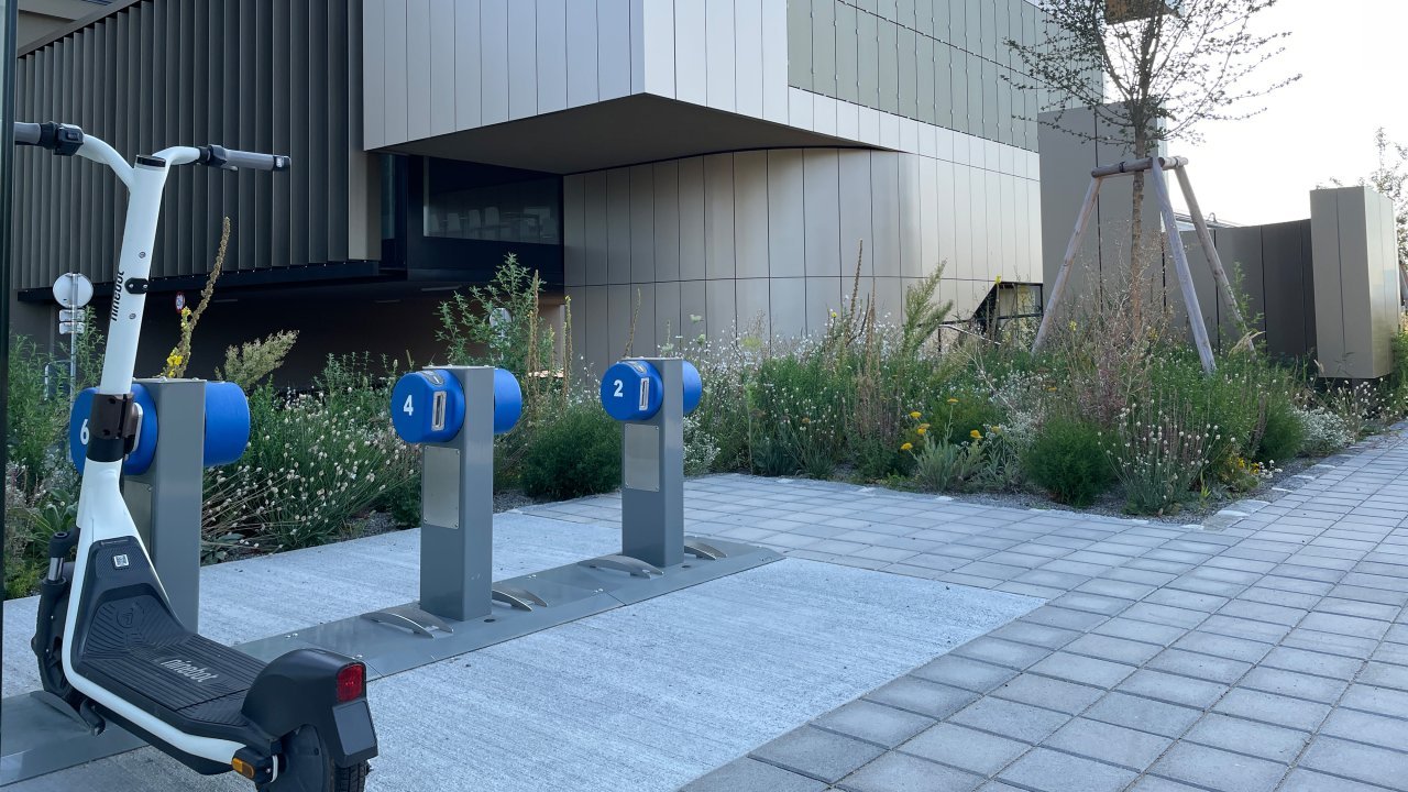 Ladestation von 3 e-Scootern vor einem modernen Firmengebäude und begrünten Eingangsbereich