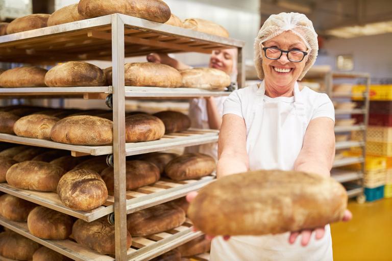 Bäckerin steht in einer Backstube mit einem Laib Brot in der Hand und Backwaren im Hintergrund