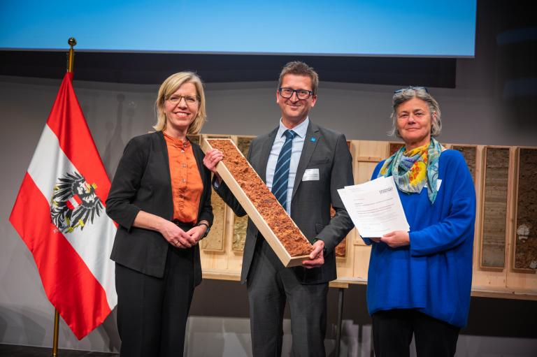Drei Personen stehen vor Bodenprofilen und einer Österreich Fahne