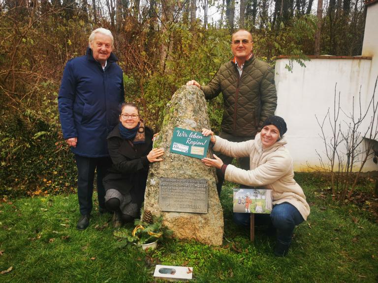 vier Personen stehen rund um einem großen Stein und präsentieren ein Schild in die Kamera.
