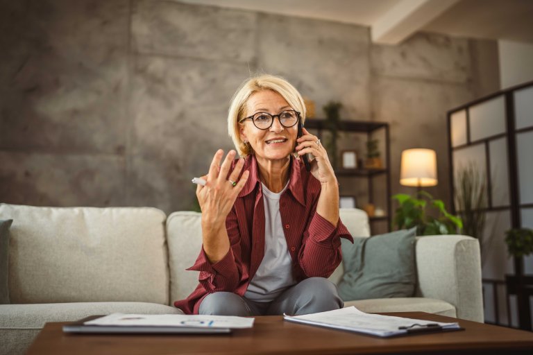 Elderly lady sits on sofa, works from home and talks on the phone