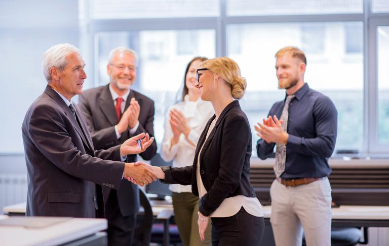 5 Personen im Business Outfit wovon sich zwei die Hände schütteln und der Rest klatscht