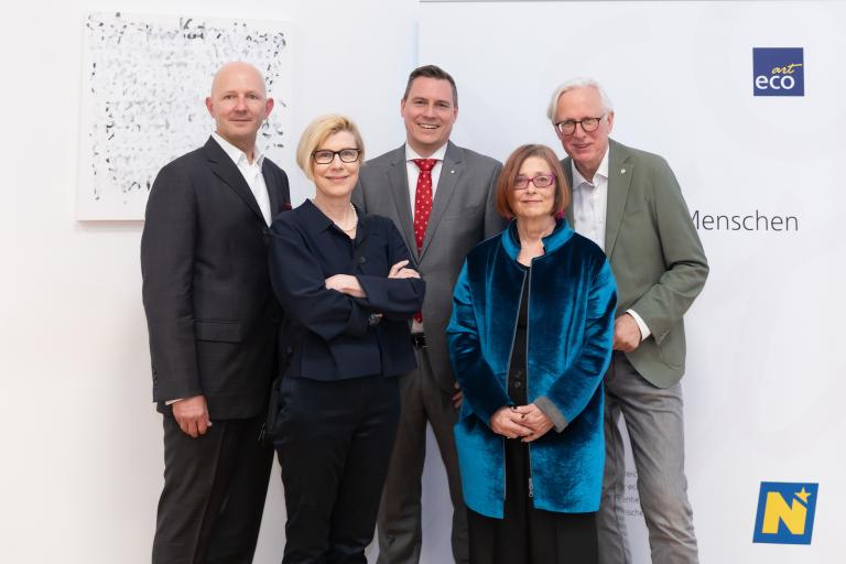 Gruppenfoto einer Vernissageneröffnung mit fünf Personen vor einem Kunstwerk und einer Logowand