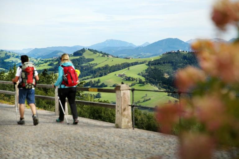 Zwei Personen wandern entlang eines Pflasterweges und im Hintergrund ist eine hügelige Landschaft. 