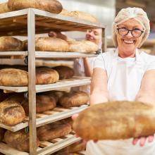 Bäckerin steht in einer Backstube mit einem Laib Brot in der Hand und Backwaren im Hintergrund