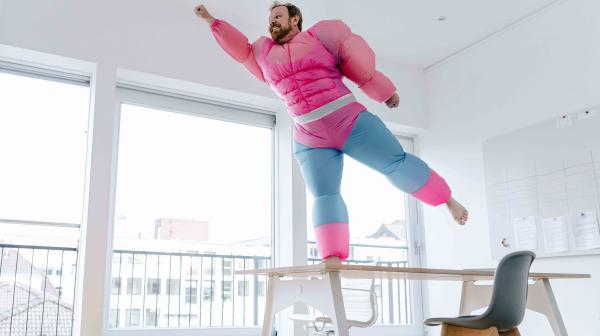 Mann in rosa Michelin-Männchenkostüm steht bloßfüssig am Tisch und streckt Arm und Bein weg