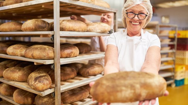 Bäckerin steht in einer Backstube mit einem Laib Brot in der Hand und Backwaren im Hintergrund