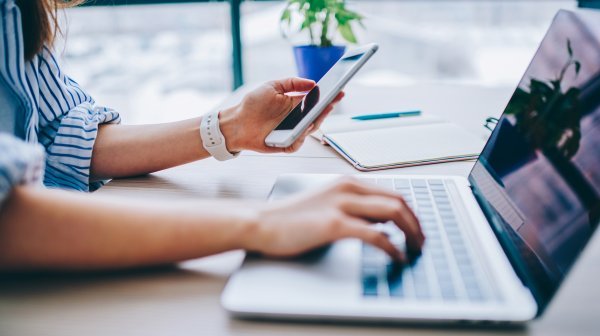 Dame tippt mit einer Hand am Computer und in der anderen Hand hält sie ihr Handy