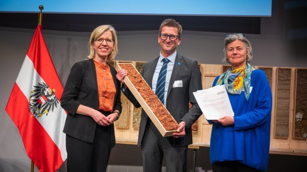 Drei Personen stehen vor Bodenprofilen und einer Österreich Fahne