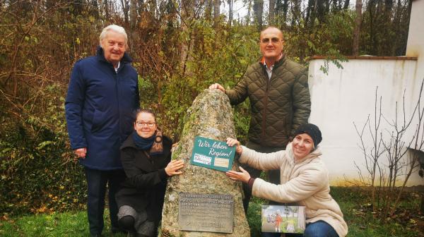 vier Personen stehen rund um einem großen Stein und präsentieren ein Schild in die Kamera.