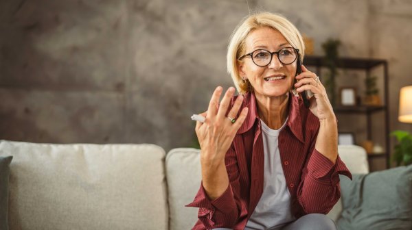 Elderly lady sits on sofa, works from home and talks on the phone
