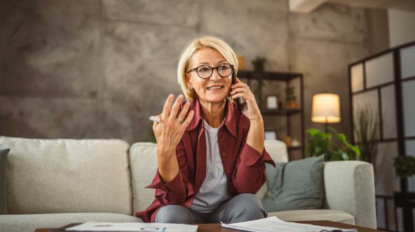 Elderly lady sits on sofa, works from home and talks on the phone