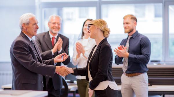5 Personen im Business Outfit wovon sich zwei die Hände schütteln und der Rest klatscht