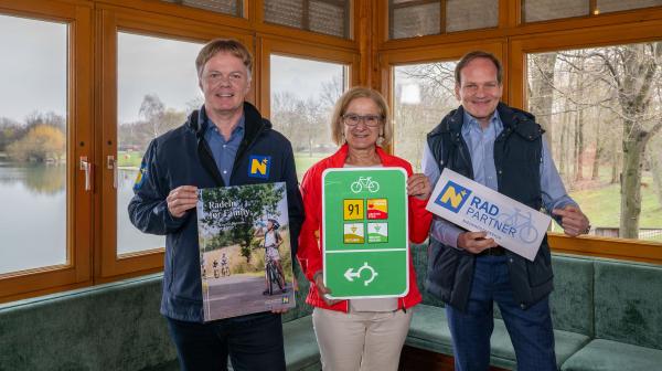 Drei Personen halten Radtafeln und Prospekt in der Hand in Zimmer mit Aussicht ins Grüne