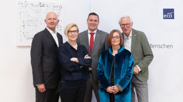 Gruppenfoto einer Vernissageneröffnung mit fünf Personen vor einem Kunstwerk und einer Logowand