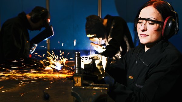 Dame arbeitet mit einem Winkelschleifer, im Hintergrund wird geschweißt