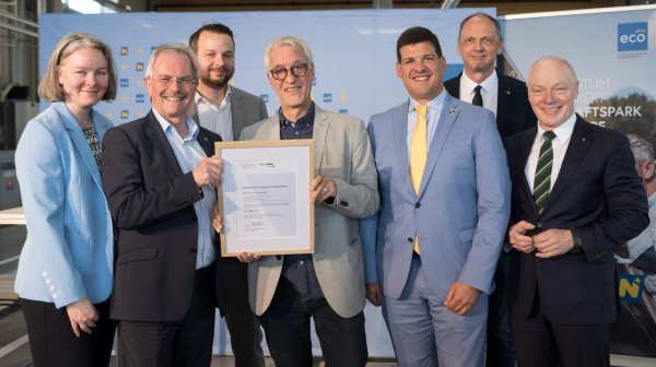 Sechs Männer und eine Frau stehen in einer Halle mit Präsentationsrückwand und halten Urkunde