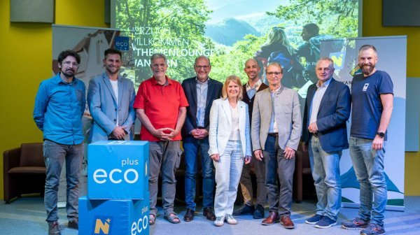 Acht männer und eine Frau stehen auf einer Bühne mit ecoplus Würfel