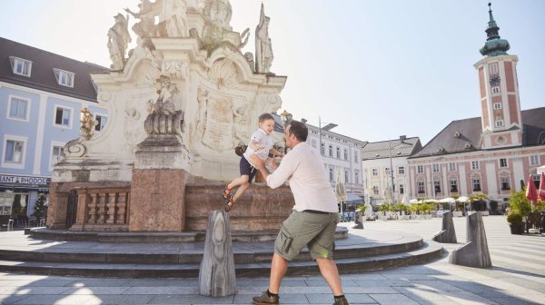 Erwachsener hebt ein Kind vor einem historischen Brunnen in einer Altstadt mit Kirchturm im Hintergrund hoch.