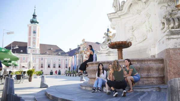 Menschen sitzen an einem Brunnen auf einem Platz mit historischen Gebäuden im Hintergrund.