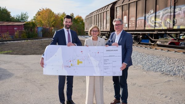 Drei Personen halten Karte mit neuen Bahngleisverlauf vor neu verlegten Bahngleisen mit 2 Bahnwaggons in die Kamera