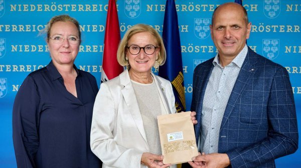 Zwei Damen mit einem Herren die Beutel mit ökonolgischen Dammmaterial vor einer Niedeösterreich-Wand mit Fahnen in der Hand halten