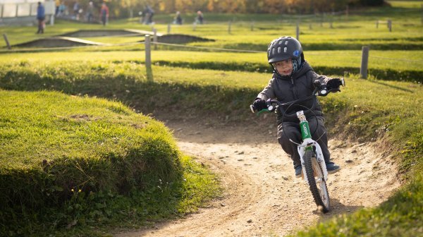 Kleiner Bub mit Helm und Wintergewand auf einem kleinen Fahrrad in einem Mini-Bikepark unterwegs