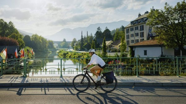 Radfahrer auf Brücke über ruhigem Fluss mit Bergpanorama im Hintergrund