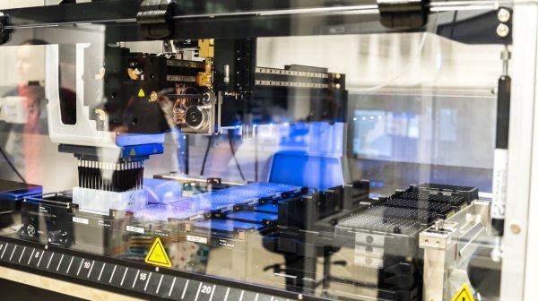 Automatisierte Laboranlage mit präzisem Pipettierroboter, der Flüssigkeiten in Mikrotiterplatten dosiert. Die Maschine ist von einem transparenten Gehäuse umgeben, mit sichtbaren Warnsymbolen und blauer LED-Beleuchtung im Inneren.