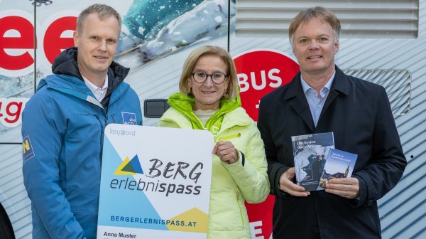 Drei Personen stehen vor einen Reisebus und halten vergrößerten Bergerlebnispass und Prospekte in die Höhe