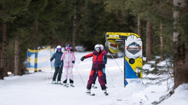 Kinder fahren auf einer schneebedeckten Waldpiste mit FunSlope-Elementen Ski