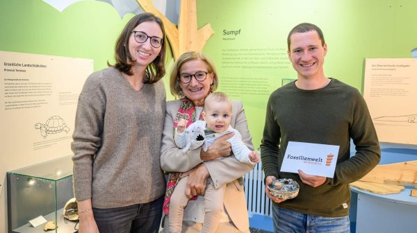 Drei Erwachsene und ein Baby sind in einer Ausstellung mit naturkundlichen Tafeln und Vitrinen. Eine Person hält ein Schild mit der Aufschrift ‚Fossilienwelt‘ und eine Schale mit Fossilien. 