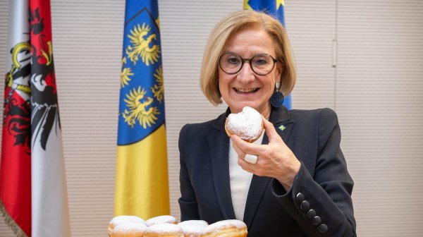 Landeshauptfrau hält ein Tablett mit Krapfen und hält einen einzelnen Krapfen in der Hand, im Hintergrund stehen eine österreichische, eine niederösterreichische und eine europäische Fahne.