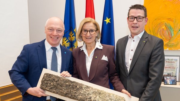 Drei Personen stehen vor drei Fahnen und halten gemeinsam eine lange, holzartige Platte in den Händen.