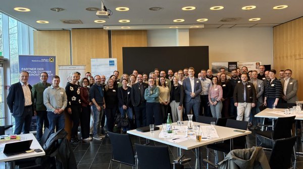 Große Gruppe von Menschen steht in einem modernen Konferenzraum für ein Gruppenfoto zusammen. Im Vordergrund befinden sich mehrere Tische mit Unterlagen, Getränkeflaschen und Stühlen.