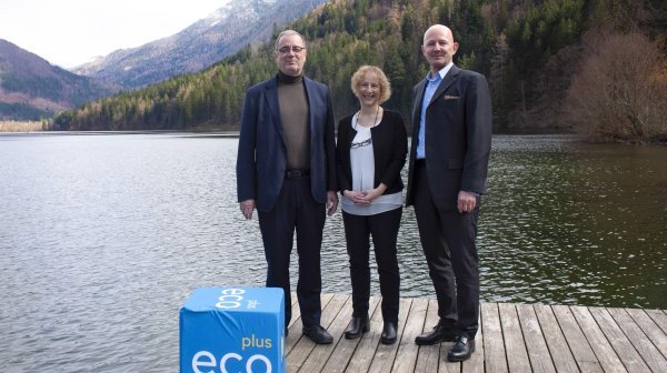 Drei Personen stehen auf einem Holzsteg an einem ruhigen See, umgeben von bewaldeten Bergen.