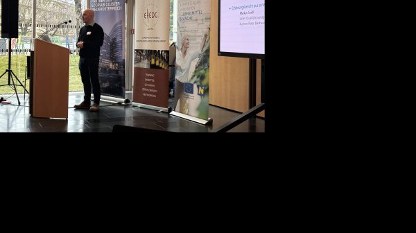 Person steht an einem Rednerpult in einem hellen Konferenzraum mit großen Glasfenstern. Hinter der Person stehen mehrere Roll-up-Banner verschiedener Organisationen. Rechts im Bild ist eine große Leinwand.