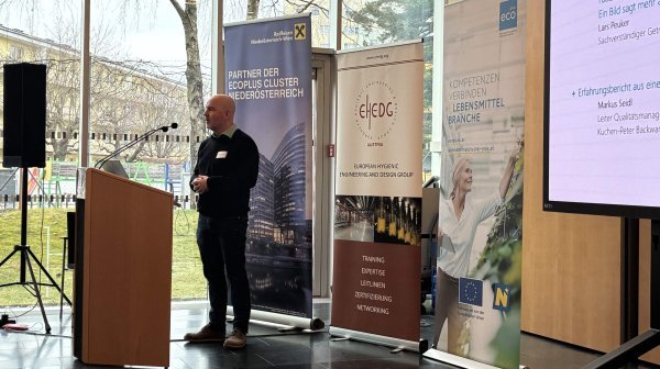 Person steht an einem Rednerpult in einem hellen Konferenzraum mit großen Glasfenstern. Hinter der Person stehen mehrere Roll-up-Banner verschiedener Organisationen. Rechts im Bild ist eine große Leinwand.